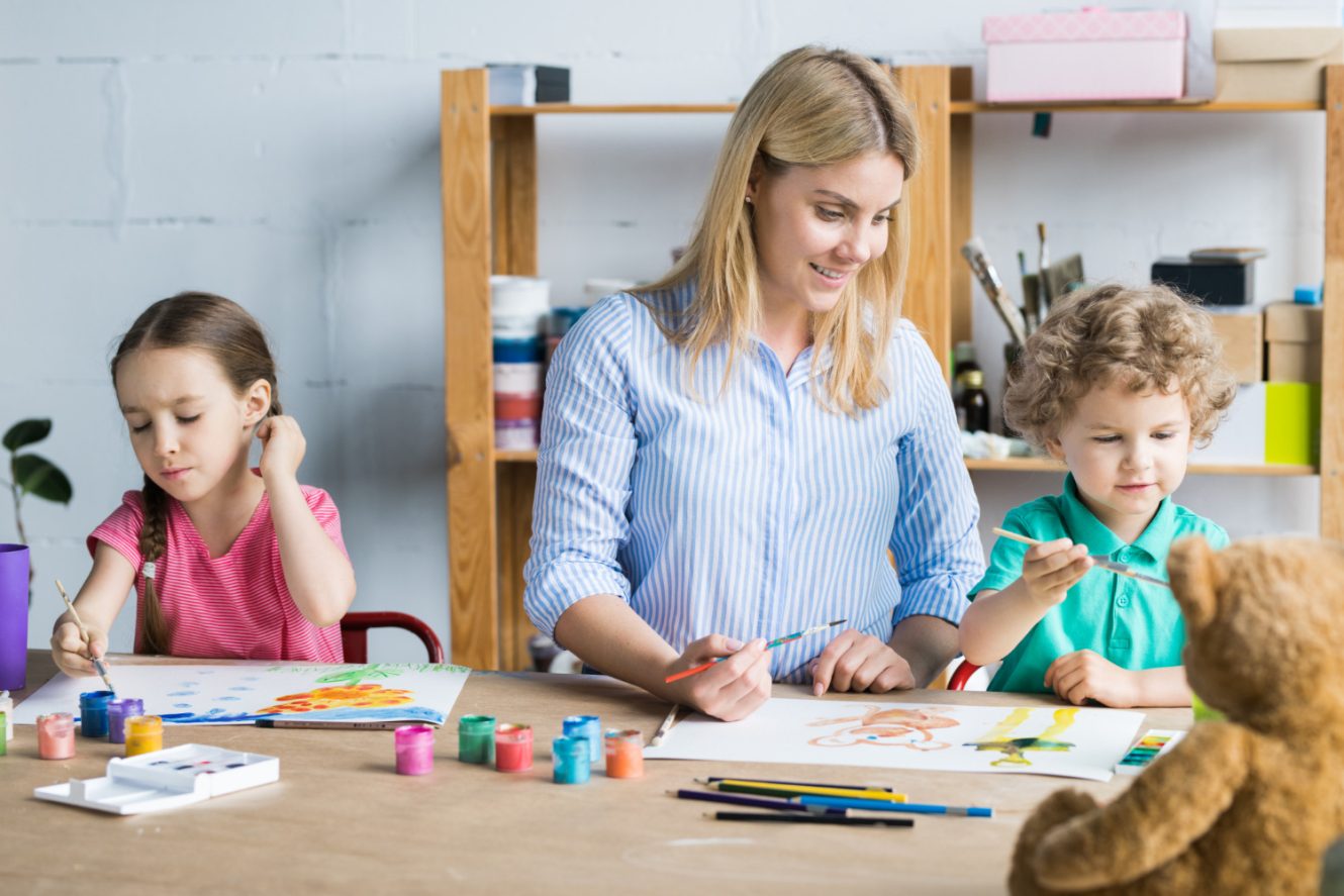 Young teacher in blue shirt helping kids draw on a paper on a wooden table